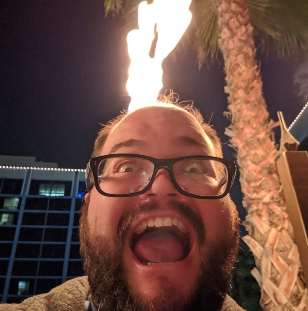 A close-up of a heavy-set, handsome bearded man with glasses, screaming into the camera. A torch right behind his head gives the illusion his head is on fire.