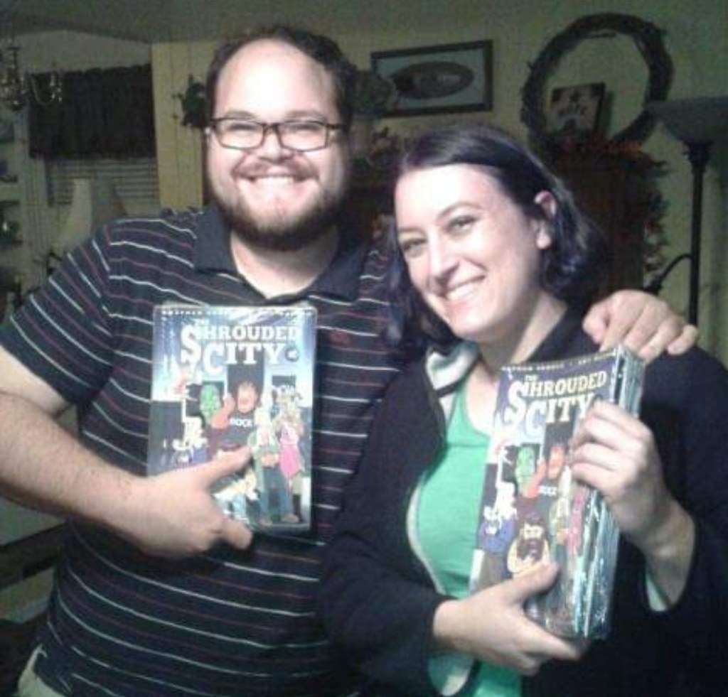 Nathan standing with Amy Watson (female, dark hair), both holding copies of their comic "The Shrouded City"