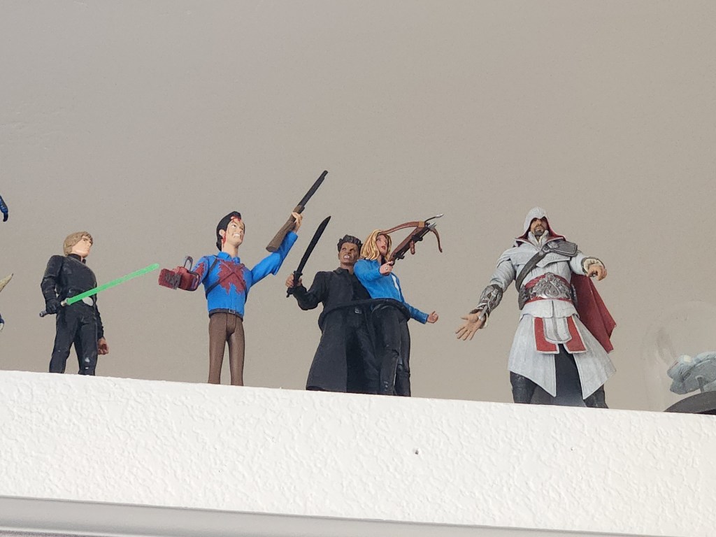 A photo from my office showing a Buffy and Angel action figure high up on a shelf, wielding weapons. Luke Skywalker, Ash Williams, and Ezio de Audiotre di Firenze are next to them.